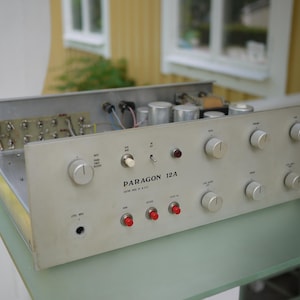 Könnte beinhalten: Ein silberner Paragon 12A Audio-Vorverstärker mit Metallgehäuse und freiliegenden internen Komponenten. Die Frontplatte verfügt über mehrere runde Drehregler, drei rote Drucktastenschalter, einen Kippschalter und eine Eingangsbuchse.