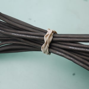 Puede incluir: Conjunto de cables de audio negros, sujetos con una cinta beige. Los cables están etiquetados con texto blanco: "Arson Audio Black Edition SC1". El fondo es de color verde azulado claro.