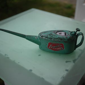 May include: A vintage teal metal oil can with a long, slender spout and a curved handle. The body features a weathered finish and a red and white label that reads Made in England Braime of Leeds. A small metal pump mechanism sits on top.