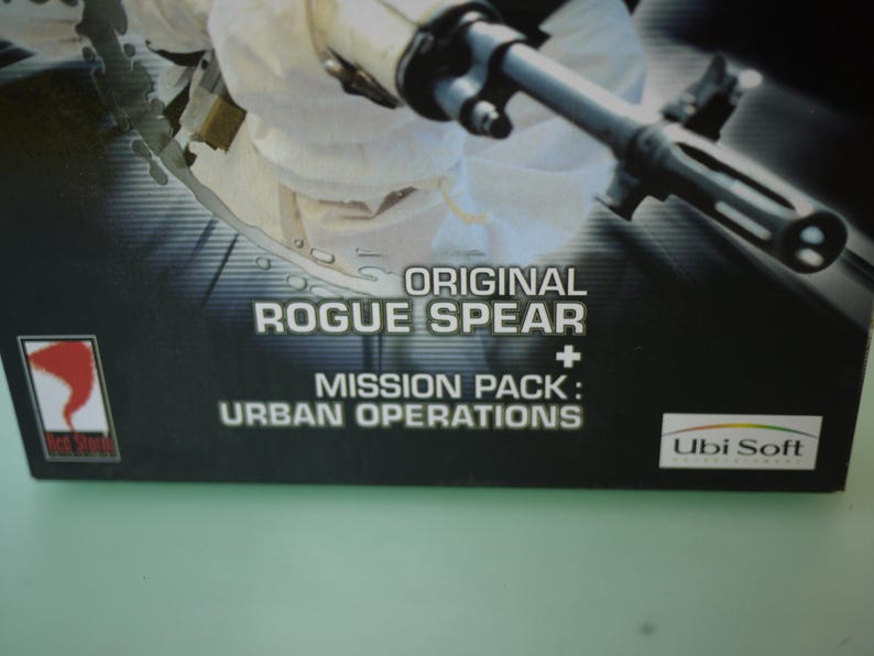 Puede incluir: Primer plano de una caja de juego con el texto "ORIGINAL ROGUE SPEAR + MISSION PACK: URBAN OPERATIONS". La caja tiene un fondo oscuro con una imagen parcial de una persona con uniforme blanco sosteniendo un arma.