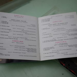 May include: An open booklet with text in Swedish, likely a program or libretto. The pages are white with black text, and the booklet is lying on a light green surface. A cassette tape is partially visible in the background.