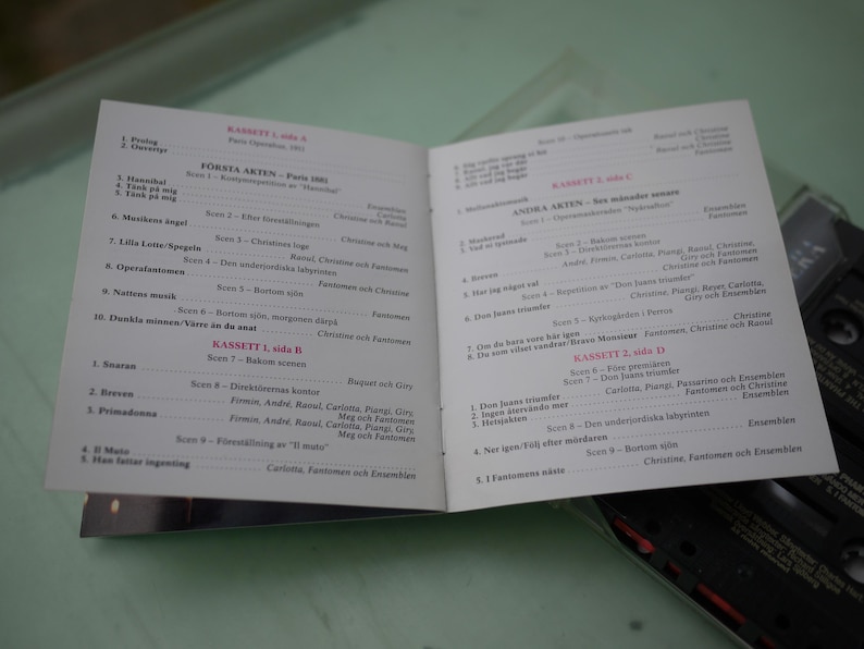 May include: An open booklet with text in Swedish, likely a program or libretto. The pages are white with black text, and the booklet is lying on a light green surface. A cassette tape is partially visible in the background.