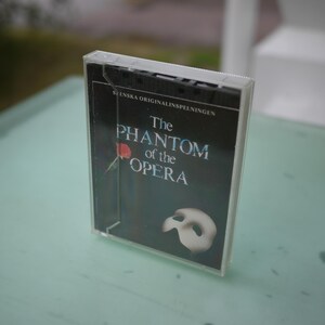 May include: A cassette tape case featuring the title "The Phantom of the Opera" in white and blue text on a black background. A red rose and a white mask are also visible. The case is clear plastic.