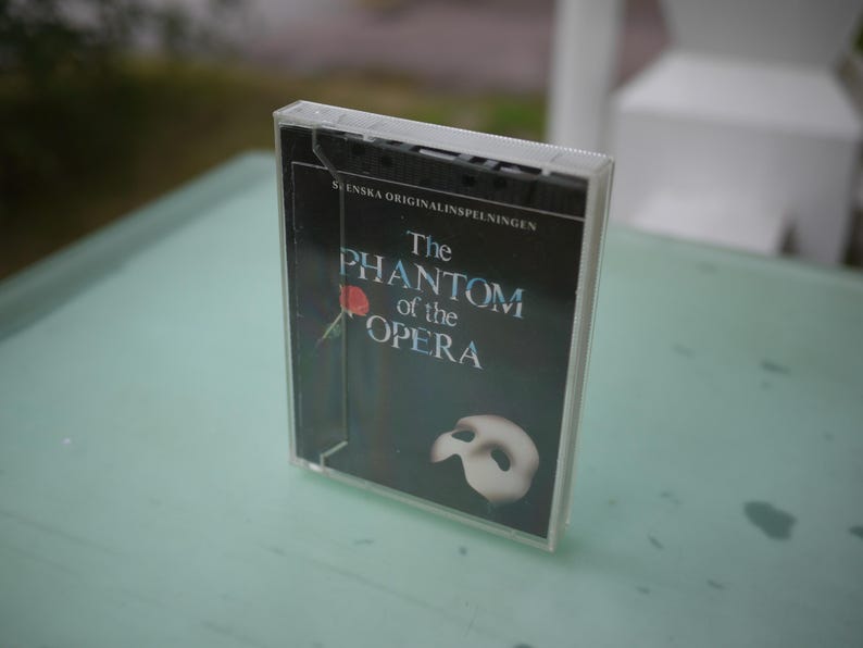 May include: A cassette tape case featuring the title "The Phantom of the Opera" in white and blue text on a black background. A red rose and a white mask are also visible. The case is clear plastic.