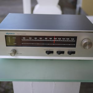 May include: A silver and black Dynaco FM tuner featuring a horizontal frequency display with a red indicator needle, a volume knob, a tuning knob, and three black toggle switches labelled Mono/Stereo, Filter, and Off/Mute on the front panel.