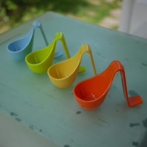 Könnte beinhalten: Vier einzelne Küchenschöpflöffel aus Kunststoff, die in einer diagonalen Reihe angeordnet sind. Jeder Schöpflöffel hat eine leuchtende Farbe: Hellblau, Limettengrün, Gelb und Orange. Das Design umfasst eine tiefe Schale und einen langen Griff.