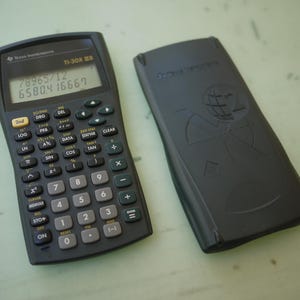 Puede incluir: Una calculadora científica Texas Instruments TI-30X IIB de color negro con una pantalla digital que muestra 78965/12 y 6580.416667, con teclado numérico y botones de función, acompañada de una cubierta protectora negra a juego.