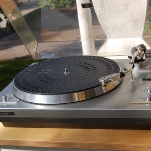Può includere: Un giradischi Garrard color argento con un piatto circolare nero dal motivo testurizzato, un braccio metallico e un coperchio antipolvere in plastica trasparente. Il dispositivo include due manopole di controllo argentate sul lato destro.