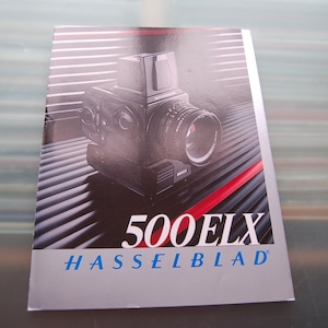 Puede incluir: Folleto con una cámara Hasselblad 500 ELX. La cámara es negra con una lente grande y un cuerpo cuadrado. El texto "500 ELX" es blanco y "HASSELBLAD" azul.