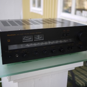 Könnte beinhalten: Ein schwarzer Onkyo MC-5000 FM-AM Stereo-Receiver. Die Frontplatte hat Knöpfe, Zifferblätter und eine Abstimmungsanzeige. Die Oberseite des Receivers hat Lüftungsschlitze. Der Receiver steht auf einem Glasregal.