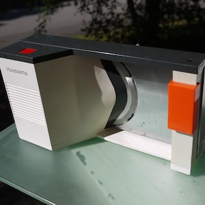 May include: A vintage Husqvarna food slicer, with a white body, a grey top, and an orange accent. The slicer has a large, circular blade and a built-in tray. The Husqvarna logo is visible on the side.