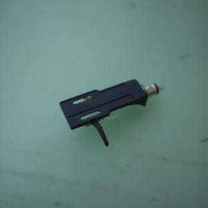 May include: A black turntable headshell featuring a rectangular body with two elongated mounting slots, a finger lift extending downwards, and a silver-toned cylindrical connector at the rear for attachment to a tonearm on a record player.