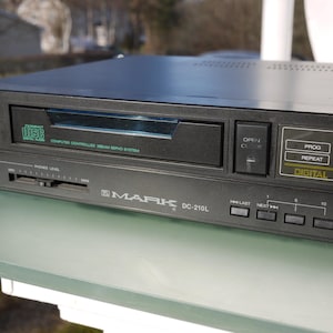 Puede incluir: Reproductor de CD Mark DC-210L negro con pantalla digital. Incluye controles de encendido/apagado, abrir/cerrar y reproducción. El reproductor de CD tiene una toma de auriculares y un control deslizante de volumen. El panel frontal tiene el texto "DIGITAL" y "COMPACT DISC DIGITAL AUDIO".