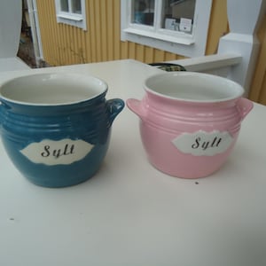 Puede incluir: Dos ollas de cerámica, una turquesa y otra rosa, cada una con interior blanco y dos asas. La palabra "Sylt" está escrita en una etiqueta blanca en la parte delantera de cada olla. Las ollas están sobre una superficie blanca.