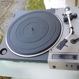 May include: A Sony turntable featuring a black circular platter, a silver metallic top panel, and a silver tonearm. The device includes a digital display screen with black control buttons and switches located on the front right corner of the unit.