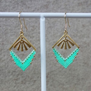 Peut inclure: Une paire de boucles d'oreilles faites à la main avec un design géométrique. Les boucles d'oreilles présentent un cadre en métal doré avec un motif de soleil et un motif tissé turquoise. Elles sont exposées sur un support blanc avec une base en bois.