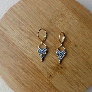Peut inclure: Une paire de boucles d'oreilles faites à la main avec un motif géométrique. Les boucles d'oreilles présentent un motif tissé bleu clair avec des accents dorés et une fermeture à levier dorée. Les boucles d'oreilles sont présentées sur une surface en bois ronde et brun clair.