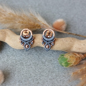 Könnte beinhalten: Ein Paar handgefertigte Ohrstecker. Jeder Ohrring hat einen kupferfarbenen Metallring mit einer zentralen Perle und aufwendigen blauen Makramee-Details. Die Ohrringe werden auf einem Stück Treibholz präsentiert.