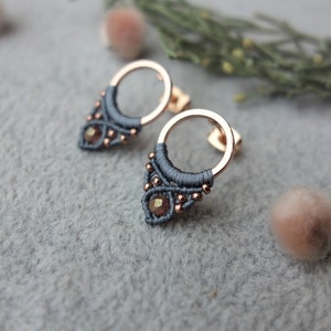 Könnte beinhalten: Ein Paar graue und roségoldene Ohrringe mit einem kleinen Kreis und einem baumelnden geometrischen Design. Die Ohrringe haben einen kleinen, dunkelbraunen Edelstein in der Mitte des geometrischen Designs.