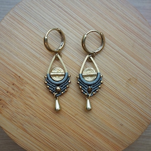 Puede incluir: Un par de pendientes de aro dorados con un colgante de borla gris y dorado. Las borlas están hechas de macramé y tienen una pequeña cuenta dorada en la parte inferior.