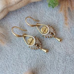 Könnte beinhalten: Ein Paar goldene Ohrringe mit weißen und braunen Perlen und einem goldenen Scheibenanhänger. Die Ohrringe liegen auf einer grauen, strukturierten Oberfläche.