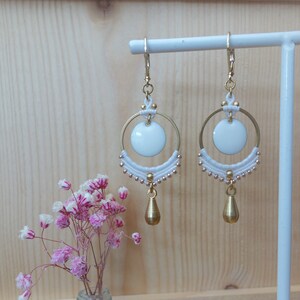 Op de afbeelding: Goudkleurige oorbellen met een cirkelvormig ontwerp. Elke oorbel heeft een witte schijf, een gouden ring en een druppelvormige hanger. De oorbellen worden gepresenteerd op een witte standaard tegen een houten achtergrond.