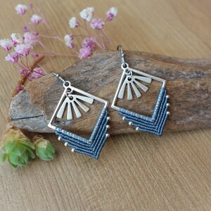 Op de afbeelding: Een paar blauwe en zilveren oorbellen met een geometrisch ontwerp. De oorbellen zijn gemaakt met een zilveren metalen frame en een blauwe koord. Het frame heeft een zonnestralenontwerp.