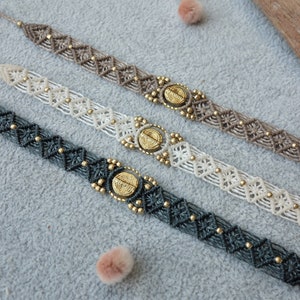 Op de afbeelding: Vier macramé armbanden in tinten grijs, wit en bruin, elk met een centraal goudkleurig medaillon en kleine gouden kralen. De armbanden worden gepresenteerd op een lichtgrijze ondergrond, die hun ingewikkelde geweven patronen laat zien.