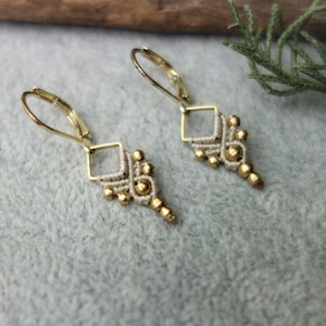 Puede incluir: Un par de pendientes de oro con un delicado diseño anudado y pequeñas cuentas doradas. Los pendientes tienen un estilo simple y elegante.