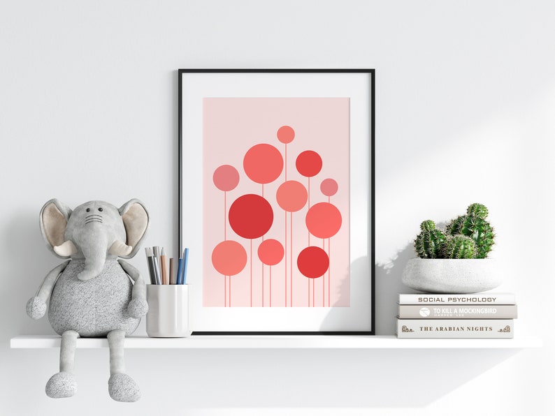May include: A framed print with a pink background and a design of red and pink circles with thin lines extending from the bottom of each circle. The print is on a white shelf with a gray elephant plush toy, a white cup with pencils, and a potted cactus on a stack of books.