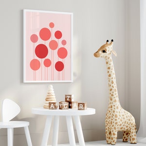 May include: A white framed print with a pink background and red circles of varying sizes. The print is hanging on a white wall. A white table with a wooden block tower and wooden alphabet blocks is in the foreground. A stuffed giraffe toy is standing on the right side of the image.