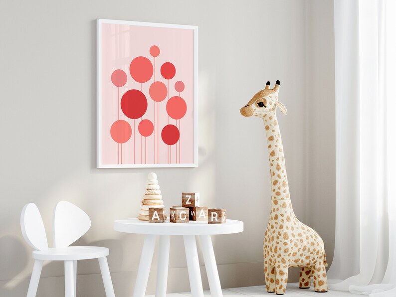 May include: A white framed print with a pink background and red circles of varying sizes. The print is hanging on a white wall. A white table with a wooden block tower and wooden alphabet blocks is in the foreground. A stuffed giraffe toy is standing on the right side of the image.