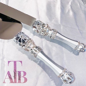May include: A silver cake knife and server set with rhinestone-encrusted handles. The handles are decorated with a floral design and have a silver finish.