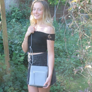 以下が含まれることがあります： 黒いオフショルダーのトップスとデニムスカートを着た女性が、ジッパー付きのグレーのクロスボディバッグを持っている。