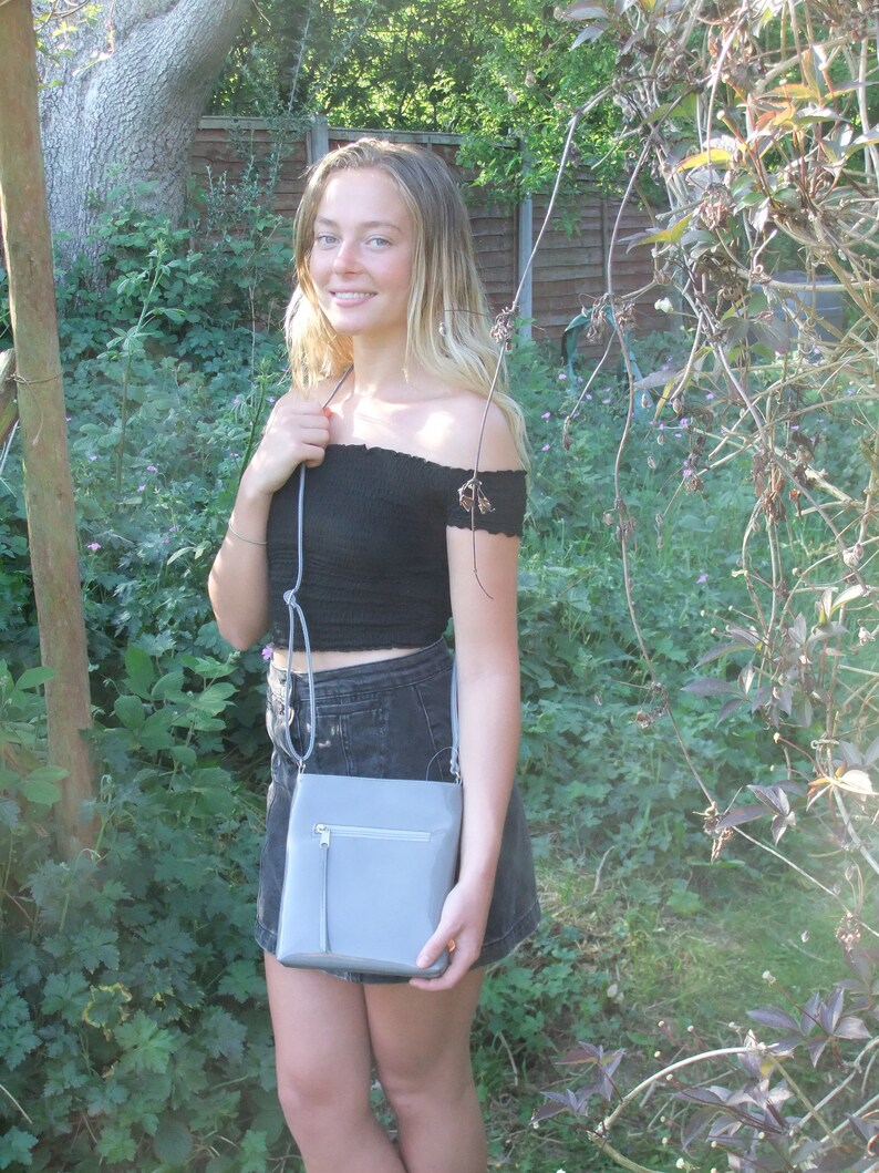 以下が含まれることがあります： 黒いオフショルダーのトップスとデニムスカートを着た女性が、ジッパー付きのグレーのクロスボディバッグを持っている。