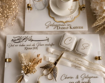 Può includere: Due vassoi rettangolari in acrilico bianco con manici dorati e trasparenti. Il vassoio superiore contiene un bicchiere e una tazzina da caffè su piattino con il testo "Süleyman in DAMAT KAHVESI". Il vassoio inferiore presenta testo in arabo, "Und wir haben euch als Paare erschaffen [QURAN 78:8]", due portaanelli con "C" e "S", un bouquet di fiori secchi, forbici dorate a forma di ali, una nappa e "Chorin & Süleyman 02.08.2026".