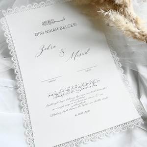 Pode incluir: Um certificado de casamento branco com uma delicada borda de renda. O documento apresenta caligrafia elegante com os nomes "Zahra & Mesud" e escrita árabe. Acentos florais secos adicionam um toque de elegância natural.