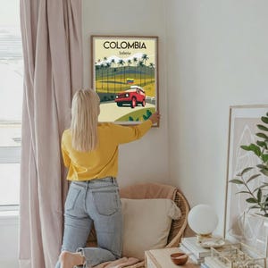 May include: A framed poster with the word "COLOMBIA" at the top, and "Salento" below it. The poster features a red jeep driving on a road, with palm trees and a Colombian flag. The poster is being hung on a white wall.