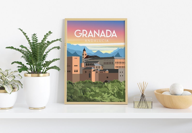 May include: A framed art print featuring a stylized illustration of Granada, Spain. The print displays the city's architecture with a sunset sky and the text "GRANADA ANDALUCIA" in white. The artwork is in a gold frame, displayed on a white shelf with plants.