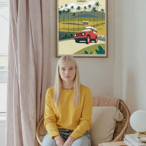 May include: A framed poster with the word "COLOMBIA" at the top, featuring a red vehicle on a road with palm trees. A person wearing a yellow sweatshirt and blue jeans sits in a wicker chair.