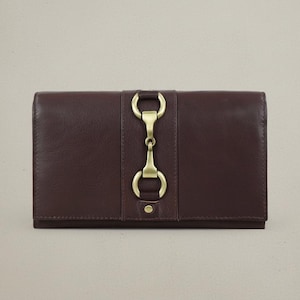 May include: A dark brown leather wallet with a brass-coloured metal clasp. The wallet is rectangular and has a vertical strap with a decorative clasp in the centre. The wallet is closed and appears to be new.