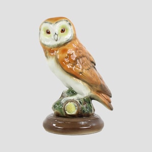 Puede incluir: Figura de búho de cerámica con cuerpo marrón y blanco, posada sobre una base de tocón de árbol verde y marrón. El búho tiene ojos grandes y expresivos y está sobre una base redonda marrón. La figura mide aproximadamente 15 cm de altura.