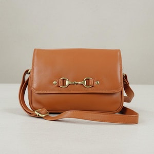 May include: A tan leather crossbody bag with a rectangular shape and a flap closure. The bag features a gold-coloured metal bit detail on the front and an adjustable shoulder strap. The bag is made of smooth leather.