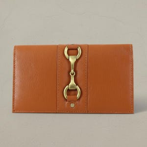 May include: A tan leather wallet with a brass clasp. The wallet is rectangular and features a central vertical brass clasp. The leather has a smooth finish. The wallet's dimensions are approximately 20 cm in length.