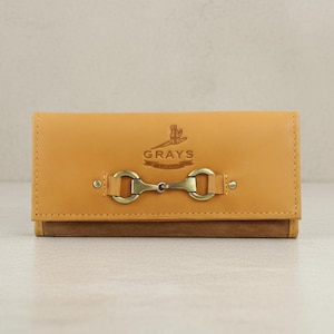May include: A tan leather wallet with a brown suede base. It has a decorative metal clasp and the word "GRAYS" printed on the front. The rectangular wallet appears to be closed. The design is simple and elegant.