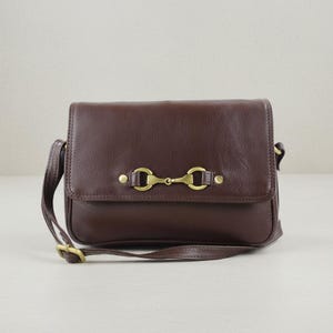 May include: A dark brown leather crossbody bag with a flap closure and an adjustable strap. The bag has a gold-coloured metal bit detail on the front. The bag is rectangular and has a classic design.