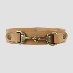 May include: A light tan leather bracelet featuring a central antique brass-toned metal bit hardware piece. The leather strap is secured on both sides of the metal bit with small, circular brass-toned metal rivets, creating a structured look.