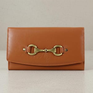 May include: A tan leather wallet with a brass-coloured metal clasp. The wallet features a flap closure and a simple design. The clasp is a horizontal bar with two circular ends. The wallet is approximately 18 cm wide.