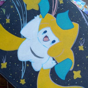 May include: A black zippered binder featuring a graphic of a white and yellow creature with blue accents, surrounded by yellow stars and light blue streaks against a dark background. The creature has a smiling face and long, flowing yellow limbs.