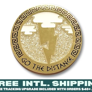 May include: Gold-coloured metal pin with a Greek key border and a lightning bolt design. The pin has a glittery texture and the words "Go the Distance" are inscribed around the edge.